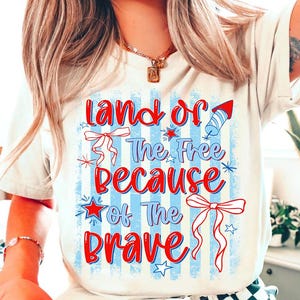 Op de afbeelding: Crèmekleurig T-shirt met de tekst "Land of the Free Because of the Brave" in rood en blauw. Het ontwerp bevat patriottische elementen zoals vuurwerk, sterren en strikken, perfect om Independence Day te vieren.