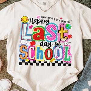 Pode incluir: Camiseta creme com um gr&aacute;fico colorido que diz "Happy Last day of School". O design inclui um rosto sorridente, uma ma&ccedil;&atilde;, um l&aacute;pis e outros elementos com tema escolar. O texto "Have fun! I love you all!" tamb&eacute;m est&aacute; inclu&iacute;do.