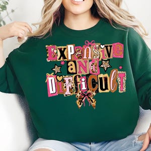 May include: A dark green sweatshirt featuring the text "Expensive and Difficult" in a colourful, block letter design. The text incorporates pink, brown, and leopard print accents, with star and bow details.