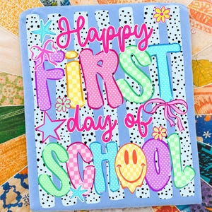 May include: A light blue rectangular fabric panel featuring the text Happy First day of SCHOOL in colorful, patterned bubble letters. The design includes black polka dots, pink bows, yellow flowers, blue starfish, and a yellow smiley face icon.