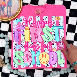 May include: A bright pink t-shirt featuring a graphic print with the text "Happy First day of SCHOOL". The design includes colorful letters with polka dot and checkerboard patterns, light blue starfish, pink bows, yellow flowers, and a yellow smiley face.