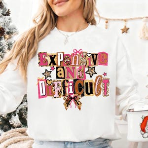 May include: White sweatshirt featuring the phrase "Expensive and Difficult" in a colourful, block letter design. The letters incorporate pink, brown, and leopard print patterns, with star and bow accents. A casual statement piece.