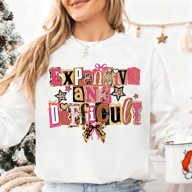 May include: White sweatshirt featuring the phrase "Expensive and Difficult" in a colourful, block letter design. The letters incorporate pink, brown, and leopard print patterns, with star and bow accents. A casual statement piece.