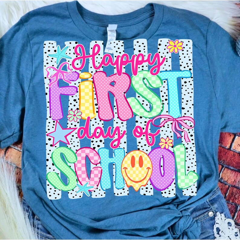 May include: A blue t-shirt featuring a graphic print with vertical black and white polka dot stripes. The text reads Happy First day of SCHOOL in colorful, patterned letters. The design includes pink bows, yellow and orange flowers, and stars.