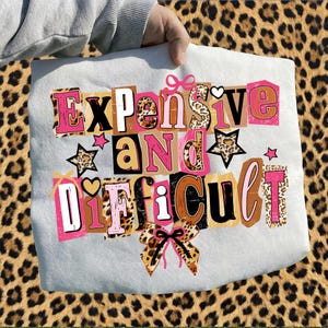 May include: A light grey tote bag featuring the phrase "Expensive and Difficult" in a collage style. The text incorporates pink, brown, and leopard print patterns. The bag is held against a leopard print background.