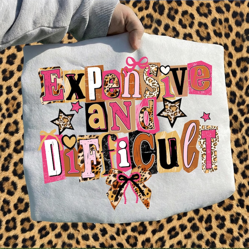 May include: A light grey tote bag featuring the phrase "Expensive and Difficult" in a collage style. The text incorporates pink, brown, and leopard print patterns. The bag is held against a leopard print background.
