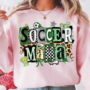 May include: Pale pink sweatshirt with the text "Soccer Mama" in a layered design. The design includes green, black, and white checkered patterns, a soccer ball, and a trophy graphic. The sweatshirt has a crew neck.