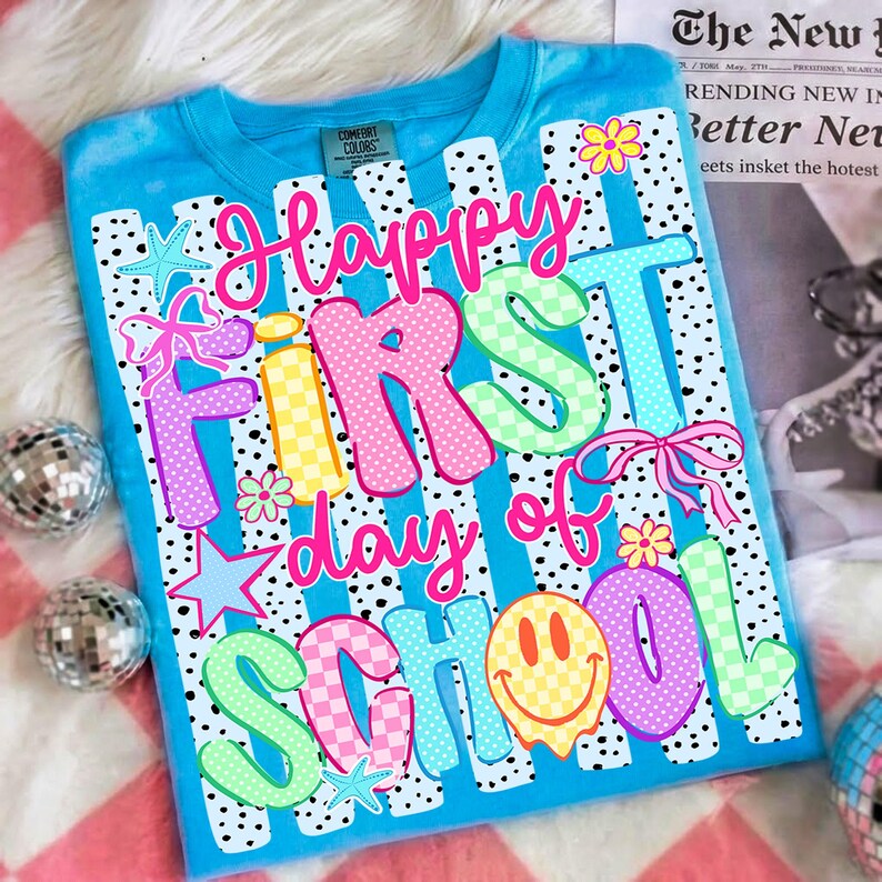 May include: A bright blue t-shirt featuring a graphic print with the text Happy First day of School in colorful, patterned bubble letters. The design includes pink bows, yellow smiley faces, blue starfish, and small flowers over a black polka dot background.