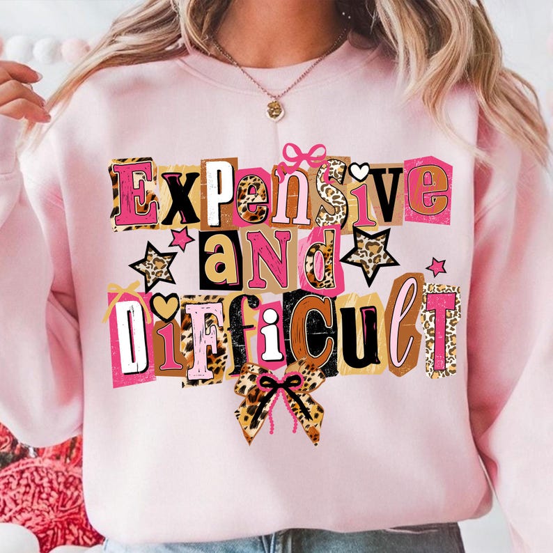 May include: Pale pink sweatshirt featuring the phrase "Expensive and Difficult" in a collage design. The text incorporates diverse fonts and patterns, including leopard print and pink. Stars and bows are used as embellishments.