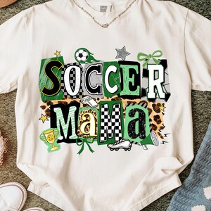 May include: Cream-coloured t-shirt featuring the words "SOCCER MAMA" in a colourful, block letter design. The design includes soccer balls, stars, and a leopard print background. The shirt is made of a soft material.