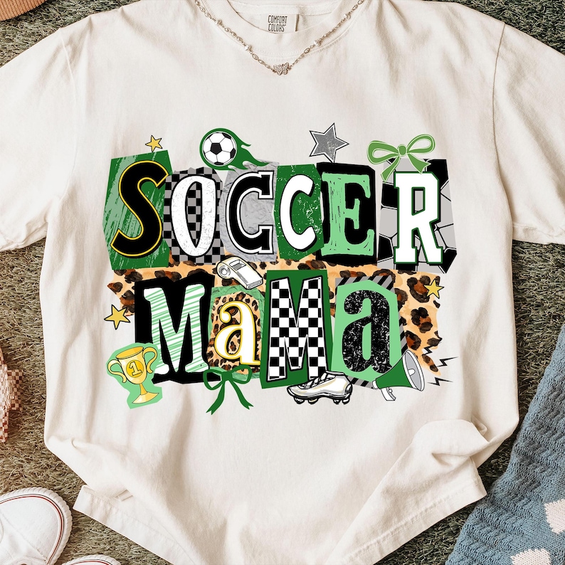 May include: Cream-coloured t-shirt featuring the words "SOCCER MAMA" in a colourful, block letter design. The design includes soccer balls, stars, and a leopard print background. The shirt is made of a soft material.