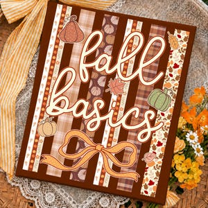 May include: A rectangular sign featuring the text fall basics in a cream script font. The background consists of vertical stripes in shades of brown, tan, and plaid, decorated with illustrations of pumpkins, autumn leaves, and a large orange bow.