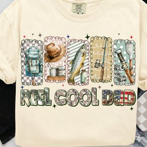 May include: Cream-colored t-shirt with a fishing-themed graphic. The design includes illustrations of fishing gear, a fish, and the text "REEL COOL DAD" in a decorative font. The shirt has a crew neck and short sleeves.