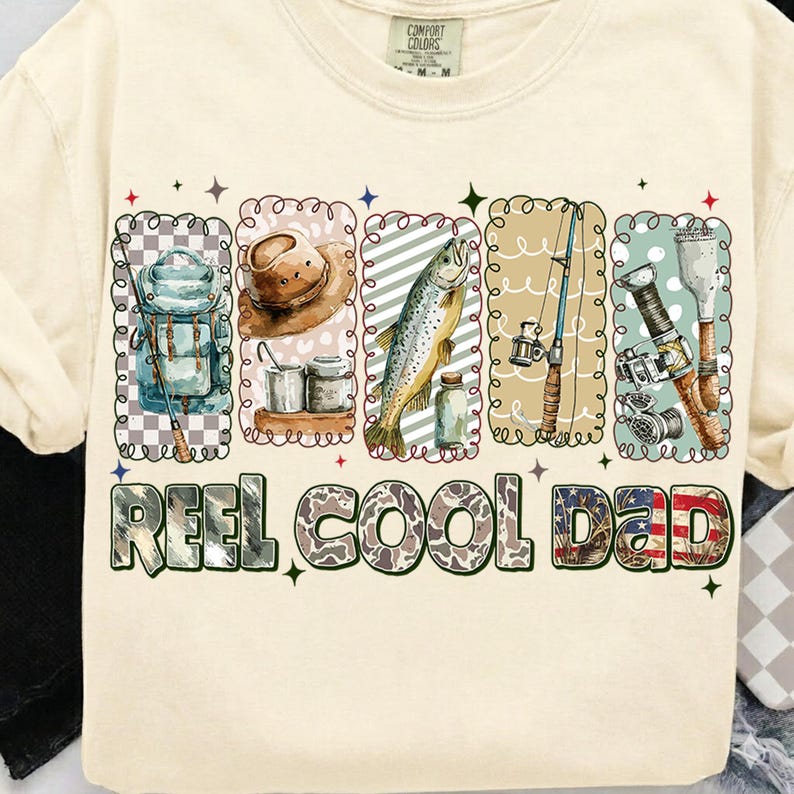 May include: Cream-colored t-shirt with a fishing-themed graphic. The design includes illustrations of fishing gear, a fish, and the text "REEL COOL DAD" in a decorative font. The shirt has a crew neck and short sleeves.