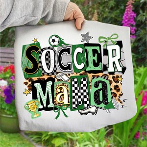 May include: A white tote bag featuring the text "SOCCER MAMA" in a layered design with soccer-themed graphics. The design incorporates soccer balls, stars, a trophy, and a football boot. The letters are in black, green, and white.