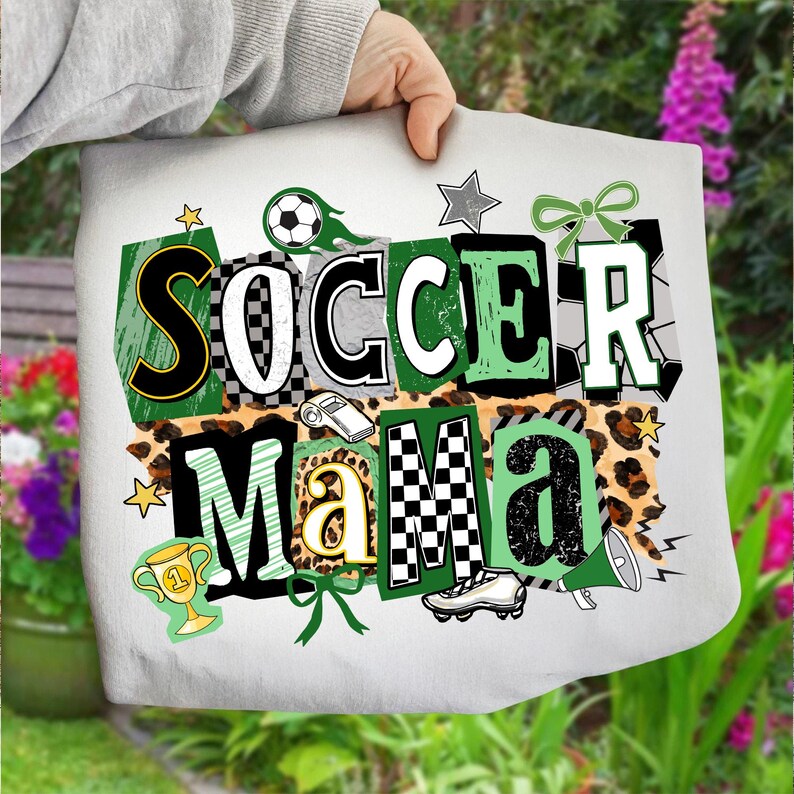 May include: A white tote bag featuring the text "SOCCER MAMA" in a layered design with soccer-themed graphics. The design incorporates soccer balls, stars, a trophy, and a football boot. The letters are in black, green, and white.