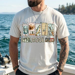 May include: Cream-colored t-shirt with a fishing-themed graphic. The graphic includes images of fishing gear, a fish, and the text "REEL COOL DAD" in a camouflage and American flag design. The shirt is worn by a person with a beard and tattoos.