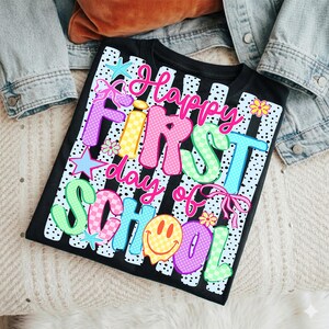 May include: A black t-shirt featuring a white vertical striped background with black polka dots. The graphic text reads "Happy First day of School" in colorful, patterned bubble letters. Accents include a yellow smiley face, stars, and flowers.