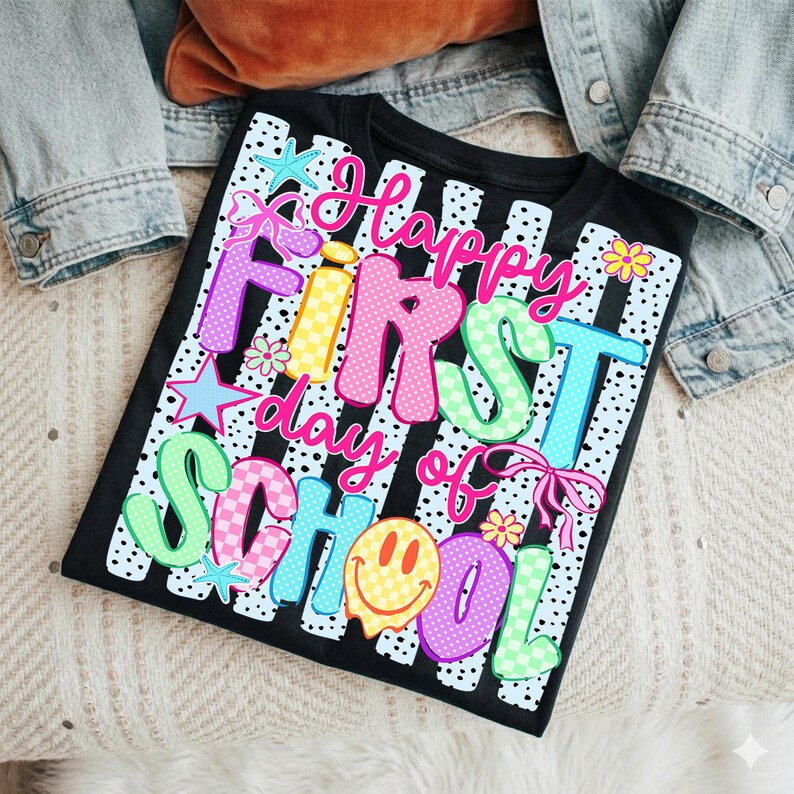 May include: A black t-shirt featuring a white vertical striped background with black polka dots. The graphic text reads "Happy First day of School" in colorful, patterned bubble letters. Accents include a yellow smiley face, stars, and flowers.