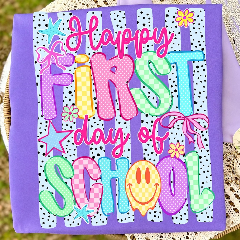 May include: A purple t-shirt featuring a graphic print with the text Happy First day of SCHOOL in colorful, patterned bubble letters. The design includes light blue vertical stripes with black dots, pink bows, yellow flowers, blue stars, and a yellow smiley face.