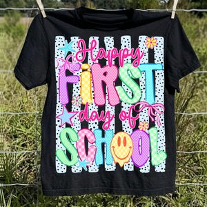 May include: A black short-sleeve t-shirt featuring a graphic print with the text Happy FIRST day of SCHOOL. The design includes vertical light blue stripes with black dots, colorful checkered and polka-dot letters, pink bows, stars, and a yellow smiley face.
