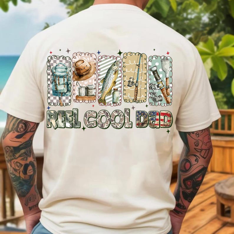 May include: Cream-colored t-shirt with a colorful graphic design featuring fishing-related images and the text "REEL COOL DAD." The design includes illustrations of fishing gear, a fish, and a hat.