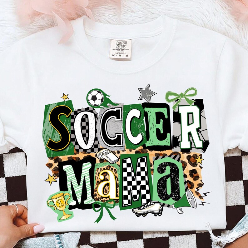 May include: White t-shirt featuring the text "SOCCER MAMA" in a vibrant graphic. The design incorporates soccer balls, stars, a trophy, and a leopard print. The shirt is a Comfort Colors brand.