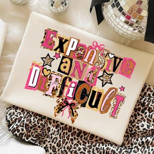 May include: A cream sweatshirt featuring the phrase "Expensive and Difficult" in a collage design. The text uses pink, brown, and leopard print elements, with star and heart embellishments. The sweatshirt is folded on leopard print fabric.
