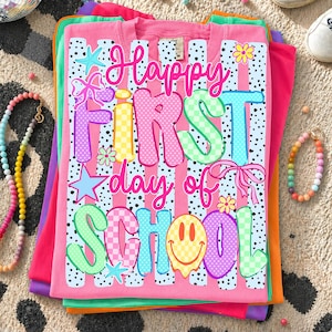 May include: A pink t-shirt featuring a graphic print with the text "Happy First day of SCHOOL" in colorful, patterned bubble letters. The design includes black polka dots, vertical stripes, stars, bows, flowers, and a yellow smiley face.