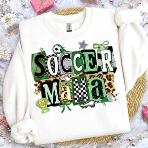 May include: White sweatshirt featuring the text "SOCCER MAMA" in a vibrant design. The design incorporates soccer balls, stars, a trophy, and a football boot. The letters are in green, black, white, and gold, set against a leopard print backdrop.