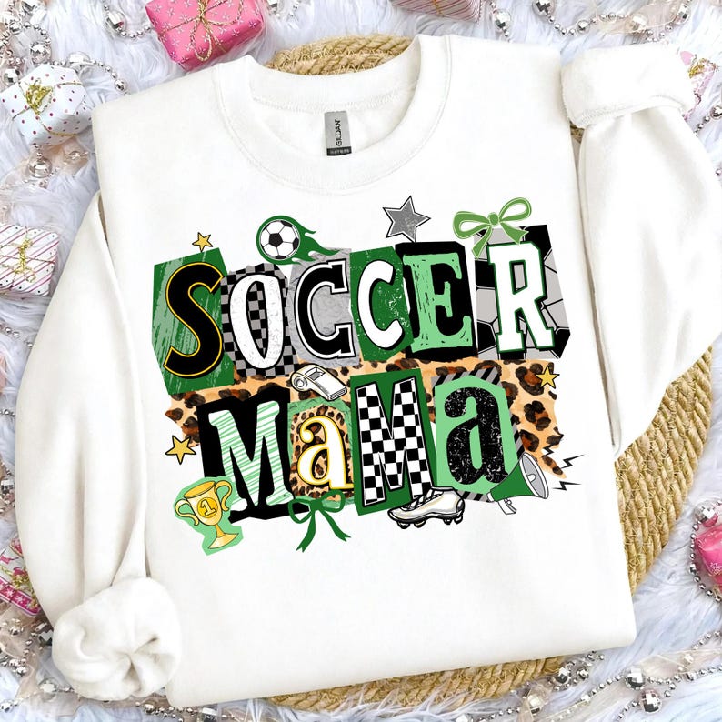 May include: White sweatshirt featuring the text "SOCCER MAMA" in a vibrant design. The design incorporates soccer balls, stars, a trophy, and a football boot. The letters are in green, black, white, and gold, set against a leopard print backdrop.