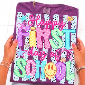 May include: A purple short-sleeved t-shirt featuring a graphic print with black and white polka-dot stripes. The text reads Happy First day of SCHOOL in colorful, patterned bubble letters, accented with starfish, flowers, bows, and a smiley face.
