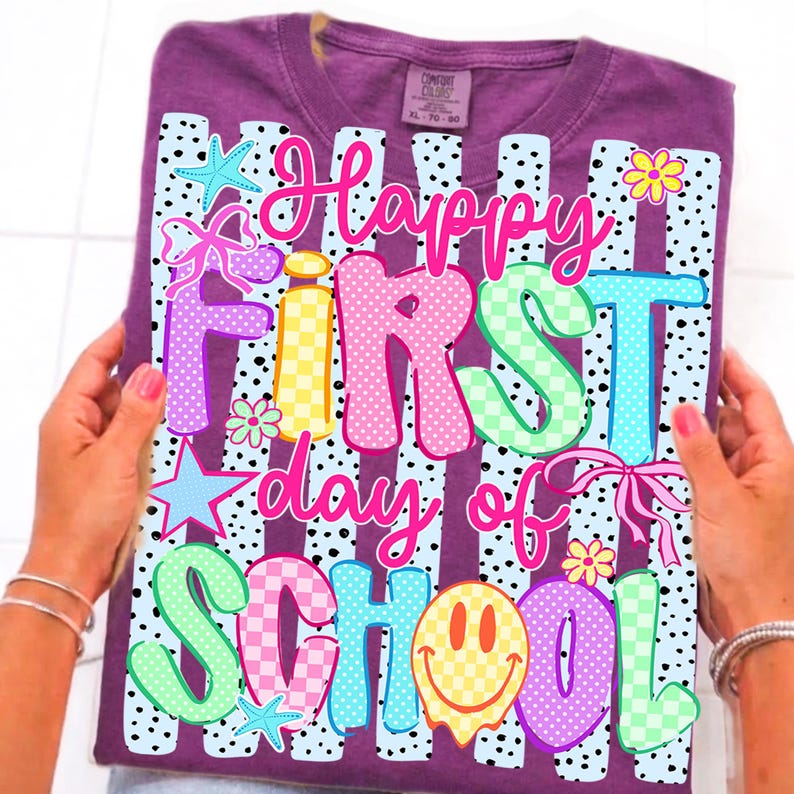 May include: A purple short-sleeved t-shirt featuring a graphic print with black and white polka-dot stripes. The text reads Happy First day of SCHOOL in colorful, patterned bubble letters, accented with starfish, flowers, bows, and a smiley face.