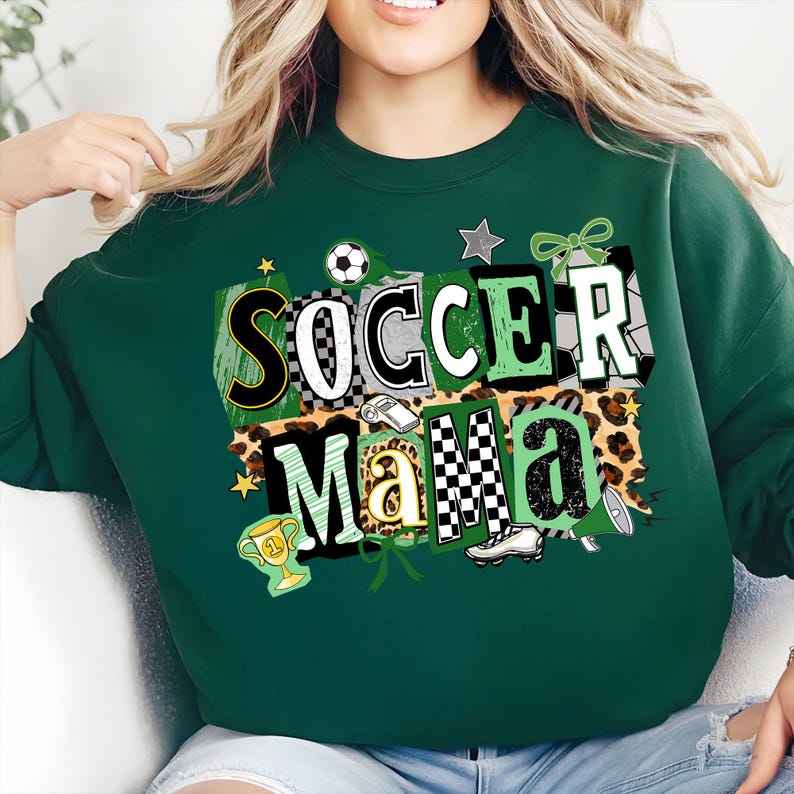 May include: A dark green sweatshirt featuring the text "Soccer Mama" in a vibrant, patchwork style. The design incorporates soccer balls, stars, and a trophy. The sweatshirt is a relaxed, casual garment.