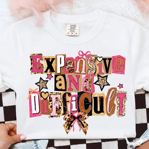 May include: White crew neck sweatshirt featuring the text "Expensive and Difficult" in a collage design. The design incorporates leopard print, pink, brown, and black colours, with star and bow accents.