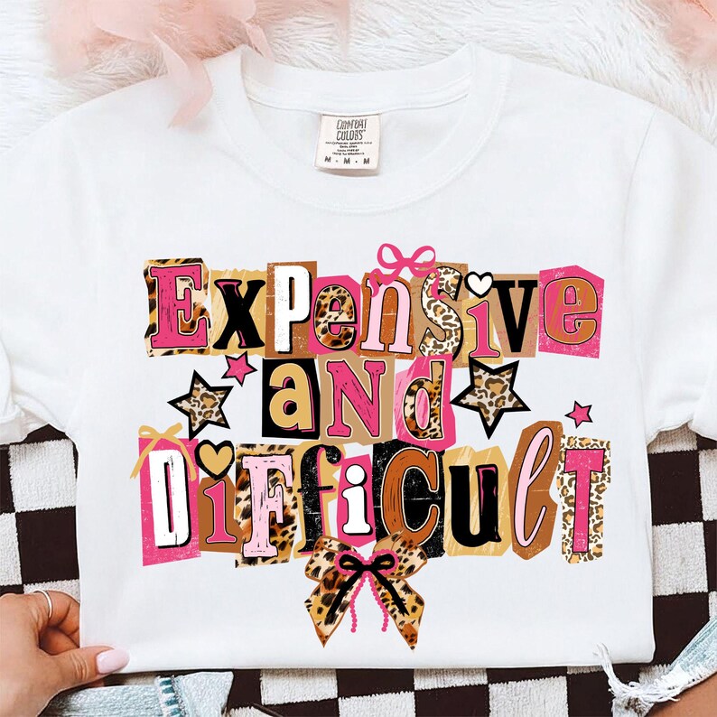 May include: White crew neck sweatshirt featuring the text "Expensive and Difficult" in a collage design. The design incorporates leopard print, pink, brown, and black colours, with star and bow accents.