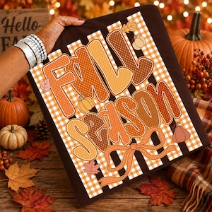 May include: A dark brown t-shirt featuring the text Fall Season in large, orange and brown polka-dot and patterned letters. The design includes a white and orange gingham background, small pumpkin illustrations, and a decorative orange ribbon bow.