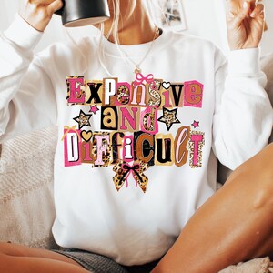 May include: White sweatshirt featuring the phrase "Expensive and Difficult" in a vibrant, block letter design. The text incorporates pink, leopard print, and brown accents, with star and bow details. A casual, statement piece.