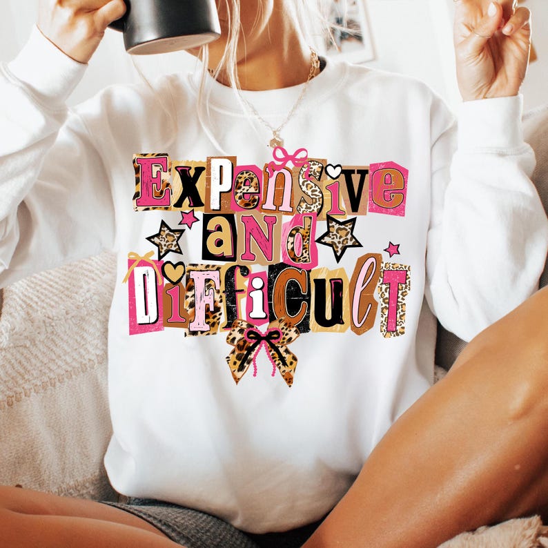 May include: White sweatshirt featuring the phrase "Expensive and Difficult" in a vibrant, block letter design. The text incorporates pink, leopard print, and brown accents, with star and bow details. A casual, statement piece.