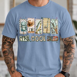 May include: Light blue t-shirt with a graphic design featuring fishing-related items such as a backpack, hat, fish, fishing rod, and tackle. The text "REEL COOL DAD" is printed below the graphics in a decorative font.