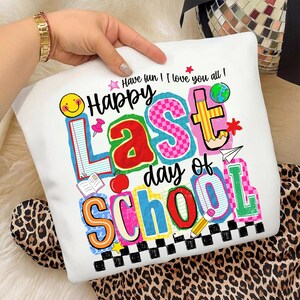 Pode incluir: Almofada branca com um gr&aacute;fico colorido que diz "Happy Last Day of School". O design inclui um smiley, um l&aacute;pis, uma ma&ccedil;&atilde; e outros elementos escolares. A almofada &eacute; segurada por uma pessoa com uma pulseira de ouro.