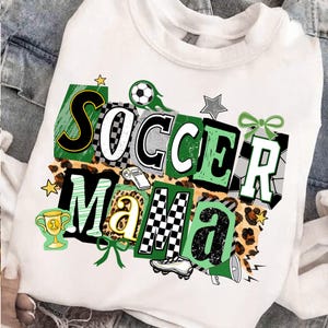 May include: White sweatshirt featuring the text "Soccer Mama" in a vibrant design. The graphic includes soccer balls, stars, a trophy, and a whistle. The letters are in different styles, with checkered and leopard print elements.