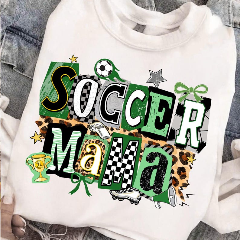 May include: White sweatshirt featuring the text "Soccer Mama" in a vibrant design. The graphic includes soccer balls, stars, a trophy, and a whistle. The letters are in different styles, with checkered and leopard print elements.