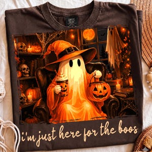 May include: A dark brown t-shirt featuring a graphic of a ghost wearing an orange witch hat, holding a pumpkin bucket and a drink. The scene includes skulls, pumpkins, and candles. The text "i'm just here for the boos" is printed in a cursive font.