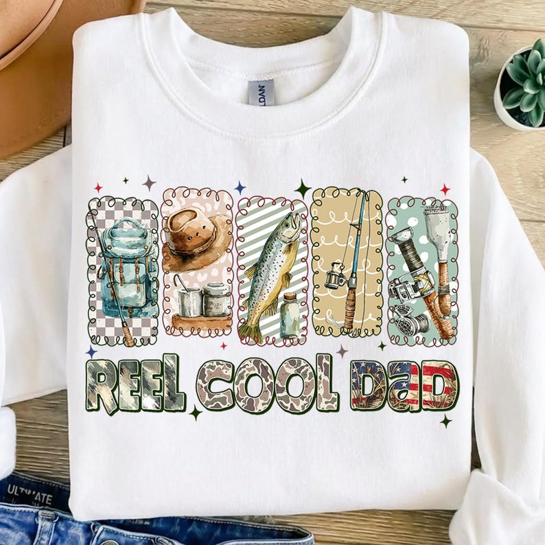 May include: White sweatshirt with a fishing-themed graphic. The design includes illustrations of fishing gear, a fish, and the text "REEL COOL DAD" in a camouflage and American flag style.