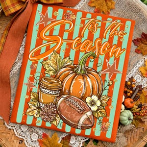 May include: A rectangular sign with orange and light blue vertical stripes featuring the text 'Tis The Season' in orange script. The design includes a large orange pumpkin, a football, a coffee cup, pinecones, and autumn leaves and stars.