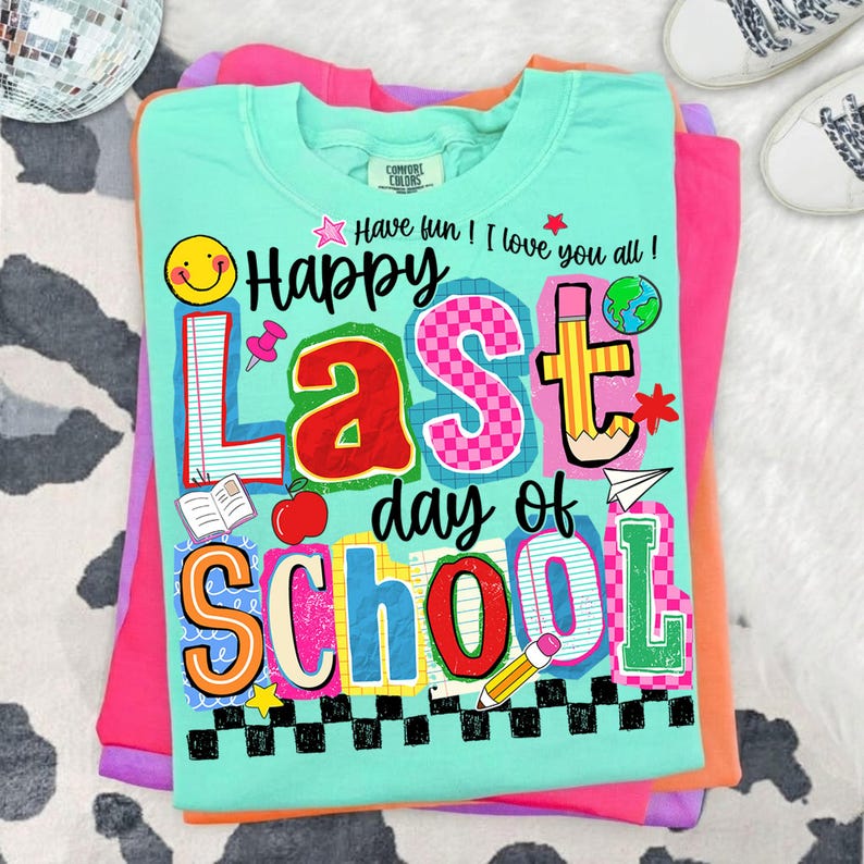 Pode incluir: Uma camiseta Comfort Colors verde claro com as palavras "Happy Last Day of School" em letras e gr&aacute;ficos coloridos. A camiseta tem um smiley, uma ma&ccedil;&atilde;, um l&aacute;pis e outras imagens com tema escolar. A camiseta est&aacute; dobrada em cima de outras camisetas coloridas.