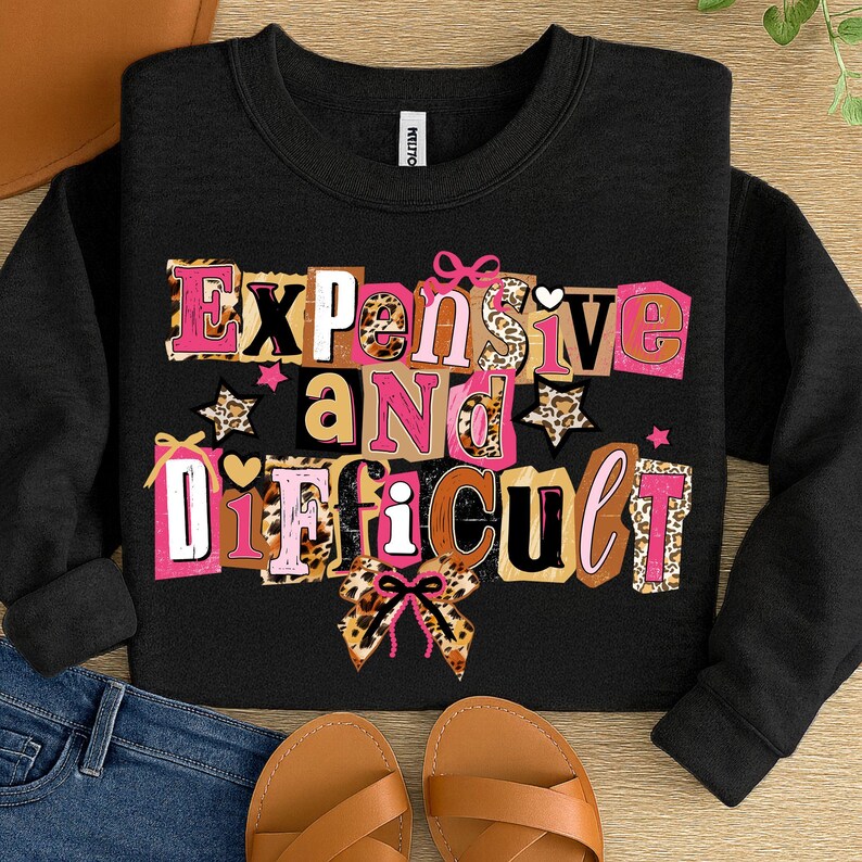 May include: Black crew neck sweatshirt featuring the phrase "Expensive and Difficult" in a colourful, block letter design. The letters incorporate pink, brown, and leopard print patterns, with star and bow accents.