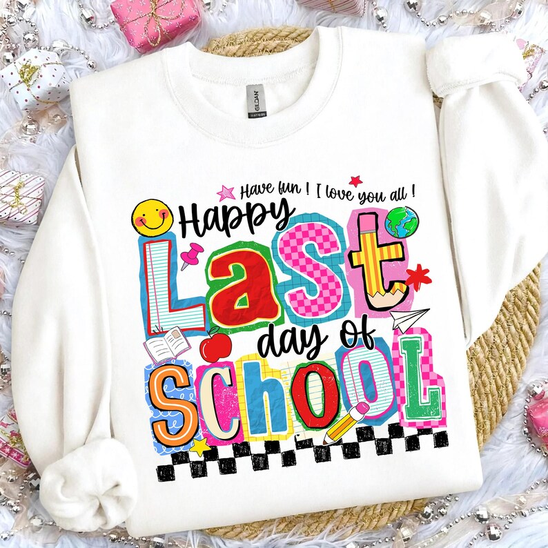Pode incluir: Camisola branca com um gr&aacute;fico colorido que diz "Happy Last Day of School". O design inclui um rosto sorridente, um globo, um l&aacute;pis e outros elementos escolares. O texto "Have fun! I love you all!" tamb&eacute;m est&aacute; inclu&iacute;do.
