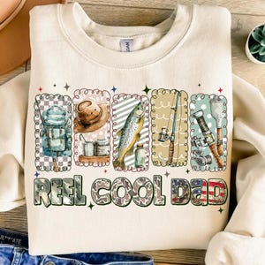 May include: Cream-colored sweatshirt with a fishing theme. The design includes illustrations of fishing gear, a fish, and the text "REEL COOL DAD" in a mix of fonts and patterns. The overall style is casual and outdoorsy.